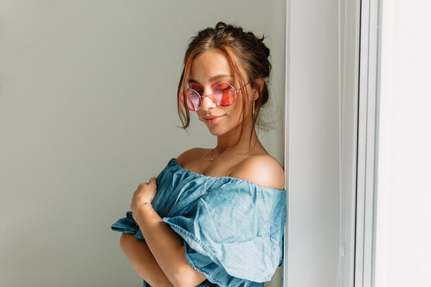 Una mujer alegre y encantadora con el pelo oscuro y gafas redondas rosas con una camiseta azul con los hombros desnudos posando cerca de la ventana a la luz del sol