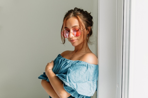 Una mujer alegre y encantadora con el pelo oscuro y gafas redondas rosas con una camiseta azul con los hombros desnudos posando cerca de la ventana a la luz del sol