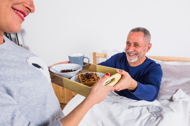 Mujer alegre envejecida que da el desayuno al hombre sonriente en el edredón en cama