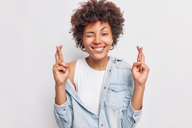 La mujer alegre y esperanzada sonríe ampliamente, mantiene los dedos cruzados, anticipa resultados positivos, usa una camisa de mezclilla, ora por la buena suerte, se opone a la pared blanca