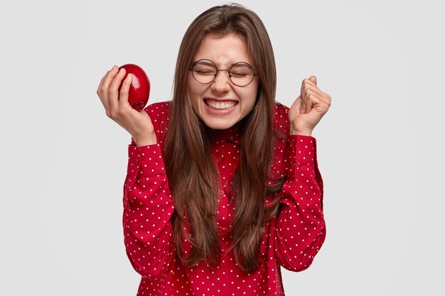 Mujer alegre con expresión complacida, aprieta el puño, tiene una sonrisa con dientes, siente placer por algo, come manzana fresca, usa anteojos redondos