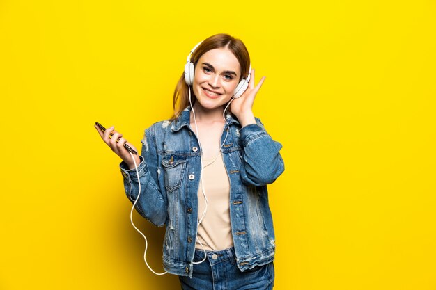 Mujer alegre feliz con auriculares escuchando música desde el estudio de teléfono inteligente disparo aislado en la pared amarilla