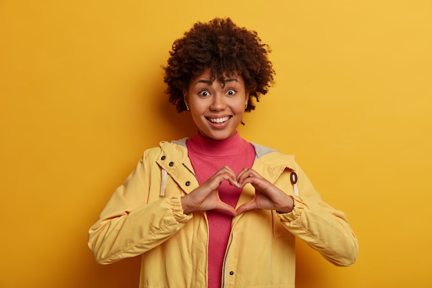 Foto gratuita la mujer alegre y femenina se confiesa enamorada, da forma al corazón sobre el pecho, expresa cuidado, dice sé mi san valentín, sonríe positivamente, encuentra el camino hacia el corazón