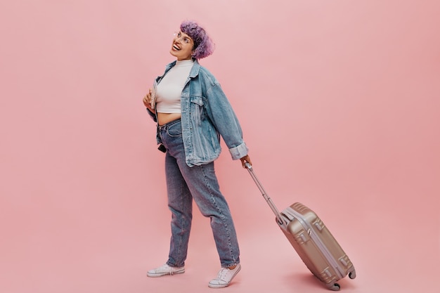 Mujer alegre con gafas y camiseta ligera y jeans ajustados pasos y sostiene la maleta. Mujer alegre con cabello morado posando.