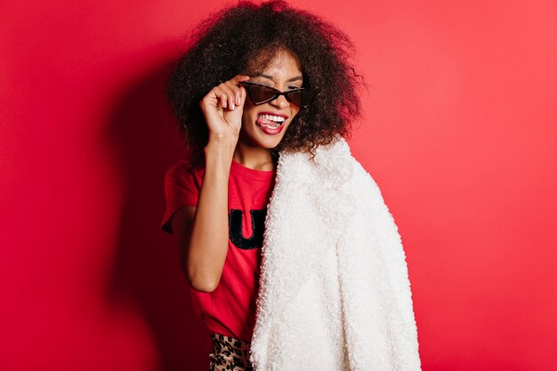 Mujer alegre en gafas de sol haciendo muecas en la pared roja