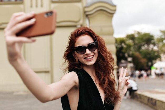 Mujer alegre con gafas de sol mostrando el signo de la paz y tomando selfie afuera