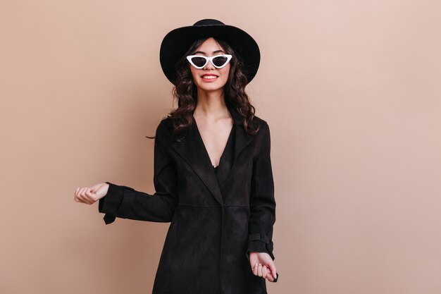 Mujer alegre con gafas de sol y sombrero mirando a cámara. Vista frontal del modelo femenino en abrigo negro.