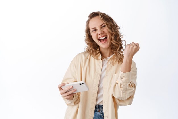 Mujer alegre ganando, logrando la meta en el teléfono móvil, sosteniendo el teléfono inteligente y gritando sí con regocijo y satisfacción, pared blanca