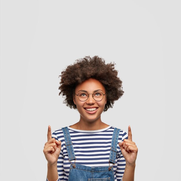 Mujer alegre con gran sonrisa, apunta hacia arriba, vestida con ropa de moda