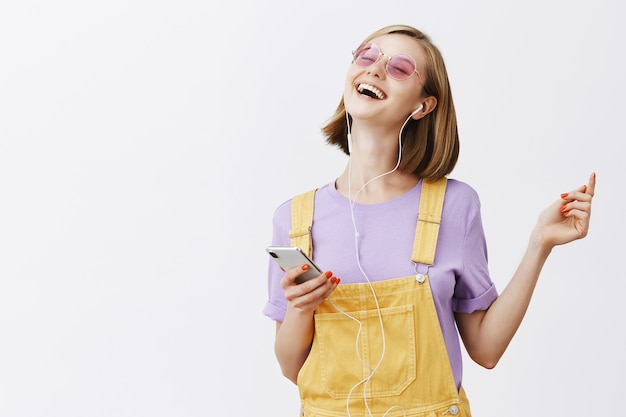 Mujer alegre guapa bailando con los ojos cerrados sin preocupaciones, sosteniendo el teléfono inteligente, escuchando música con auriculares