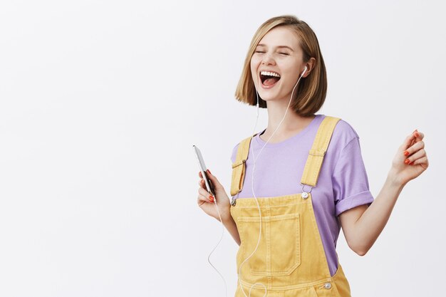 Mujer alegre guapa bailando con los ojos cerrados sin preocupaciones, sosteniendo el teléfono inteligente, escuchando música con auriculares