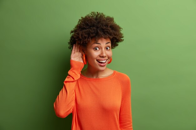 Una mujer alegre y guapa con cabello rizado mantiene la mano cerca de la oreja mientras trata de escuchar una conversación interesante, escucha atentamente a los compañeros de trabajo que hablan en privado, alegres y curiosos, se para en el interior