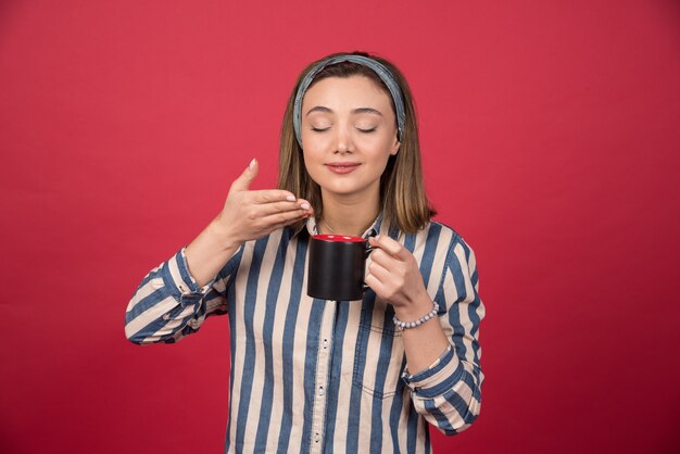 Mujer alegre huele el aroma del café en la pared roja