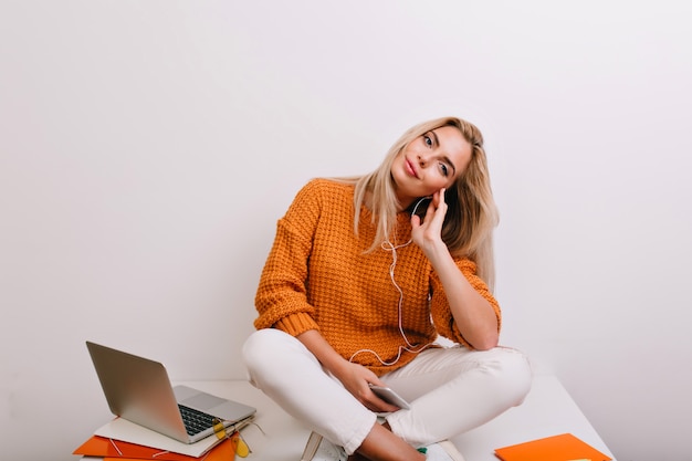 Mujer alegre en jeans blancos sentada en la mesa con auriculares y descansando después de un duro día de trabajo