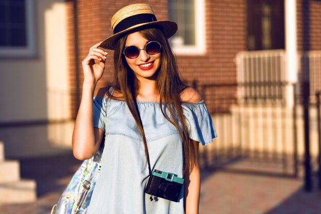 Mujer alegre joven bastante hipster posando en la calle en un día soleado, divirtiéndose solo, sombrero de ropa vintage con estilo y gafas de sol, concepto de viaje, joven fotógrafo con cámara vintage.