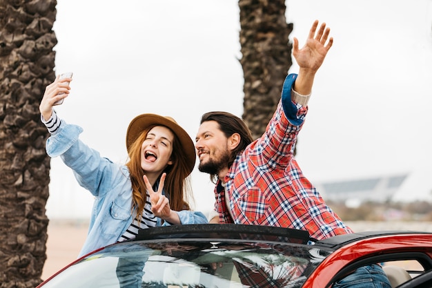 Mujer alegre joven y hombre que se inclinan hacia fuera del coche y que toman el selfie en smartphone