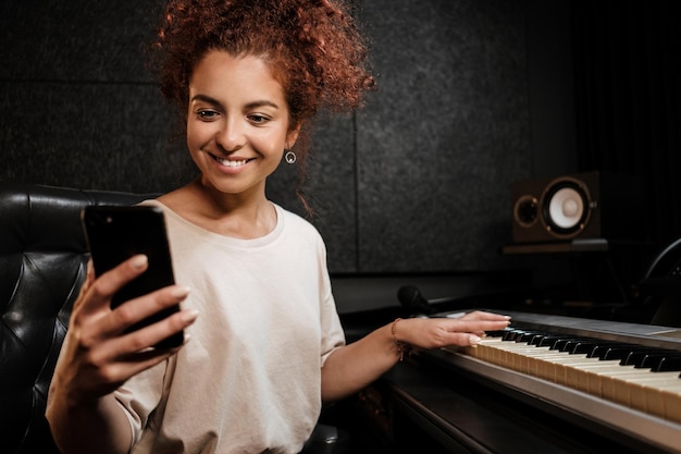 Mujer alegre joven que usa felizmente el teléfono tocando el piano eléctrico en el estudio de grabación de sonido