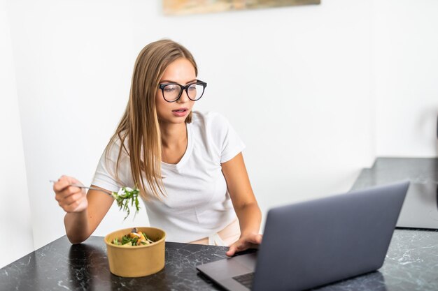 Mujer alegre joven usando computadora portátil y comiendo ensalada en la cocina