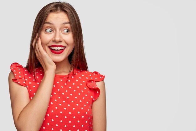 Mujer alegre con lápiz labial rojo posando contra la pared blanca