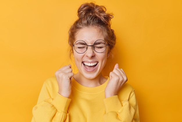 Una mujer alegre y llena de alegría con el pelo peinado celebra los vítores de la victoria y exclama de alegría mantiene los ojos cerrados vestidos casualmente tiene el pelo peinado aislado sobre un fondo amarillo vivo. yo gano