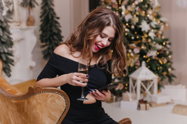 Mujer alegre con mensaje de texto de pelo largo brillante en la noche de año nuevo. Retrato interior de una chica de moda en vestido negro bebe champán y sosteniendo el teléfono cerca del árbol de Navidad.