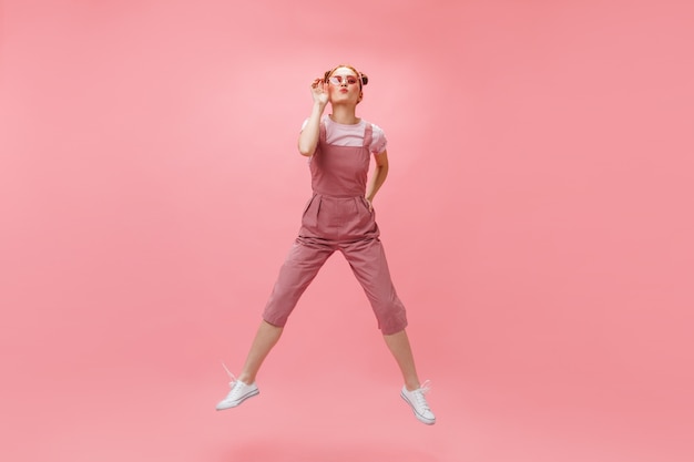 Mujer alegre en monos y camiseta blanca se pone gafas de sol y salta sobre fondo rosa.