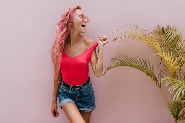 mujer alegre en pantalones cortos de mezclilla posando junto a la palmera.