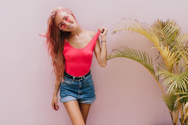 mujer alegre en pantalones cortos de mezclilla posando junto a la palmera.