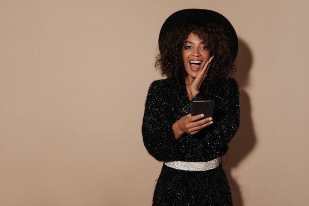 Mujer alegre con peinado morena con sombrero de moda y suéter negro con cinturón brillante mirando a cámara y sosteniendo el teléfono inteligente en la pared beige.