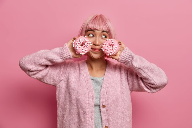 Mujer alegre con peinado rosa, sostiene dos donas glaseadas, poses