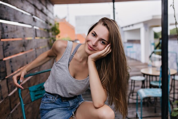 Mujer alegre con el pelo largo posando en el café de la calle con expresión de cara de ensueño. Chica de pelo oscuro en falda de mezclilla sentada en una silla en la escena urbana.