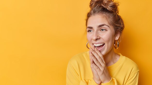 Una mujer alegre con el pelo peinado mantiene las manos juntas cerca de la boca se ríe positivamente de una broma divertida concentrada en la distancia usa aretes y un jersey aislado sobre un espacio en blanco en la pared amarilla