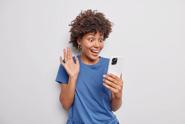 Una mujer alegre con el pelo rizado se hace selfie o hace videollamadas con gesto de saludo ha sorprendido la expresión feliz escucha buenas noticias viste una camiseta azul casual aislada sobre fondo blanco