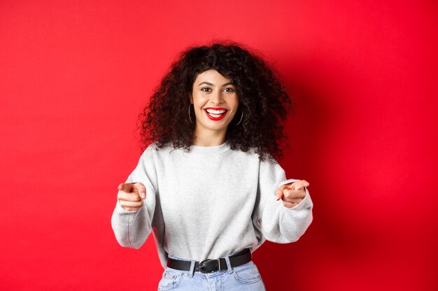 Mujer alegre con el pelo rizado que le invita a reclutar novatos señalando con el dedo a la cámara y sonriendo ...