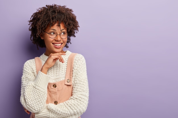 Foto gratuita la mujer alegre de piel oscura tiene una expresión pensativa, mantiene la mano debajo de la barbilla, tiene una mirada tierna y tierna, sonríe cortésmente, espera al cliente en la tienda, vestida con ropa de moda, aislada en la pared púrpura