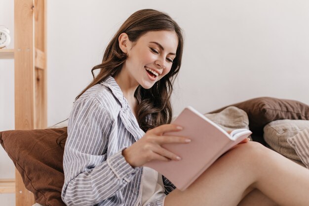 Mujer alegre en pijama de rayas azules con libro de lectura de sonrisa sentado en el sofá