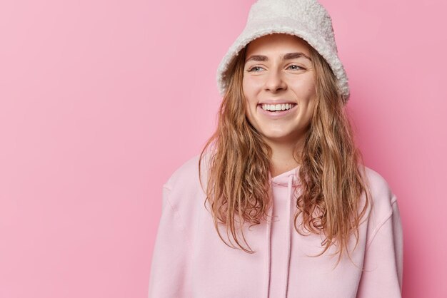 La mujer alegre y positiva sonríe ampliamente, mira hacia otro lado, se siente feliz, usa panama y sudadera con capucha, está de buen humor aislada sobre el espacio de copia en blanco de fondo rosa para su contenido promocional. Concepto de emociones