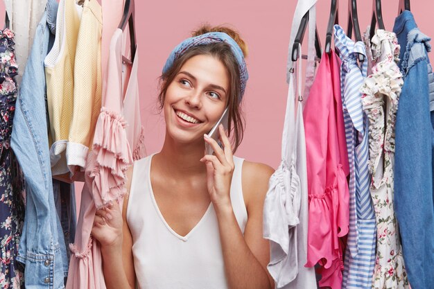 Mujer alegre positiva con una sonrisa agradable, conversando por teléfono inteligente mientras está de pie entre perchas con ropa. Feliz mujer vestida casualmente sonriendo mientras habla por teléfono móvil