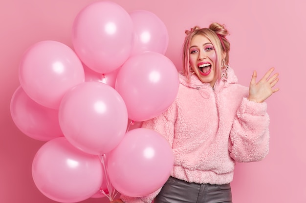 La mujer alegre y positiva tiene un maquillaje brillante y exclama que algo en voz alta mantiene la palma levantada vestida con ropa de moda sostiene un gran montón de globos en la fiesta de cumpleaños. Concepto de vacaciones y personas