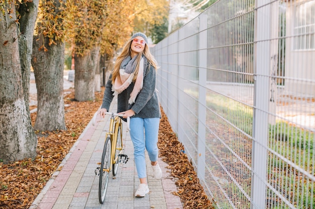 Mujer alegre que camina con la bicicleta cerca de la cerca