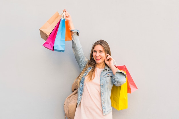 Mujer alegre que está parada con los bolsos de compras brillantes en la pared ligera