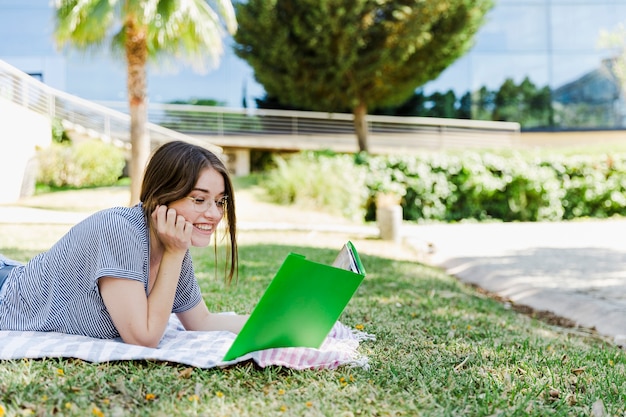 Mujer alegre que lee el libro de texto en hierba del parque
