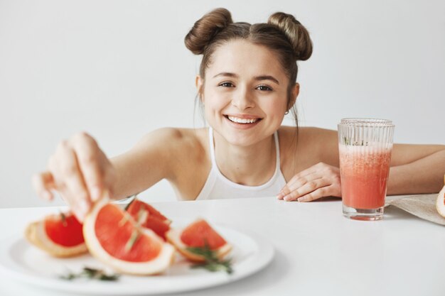 Mujer alegre que sonríe tomando la rebanada de pomelo de la placa que se sienta en la tabla sobre la pared blanca. Comida saludable y saludable.