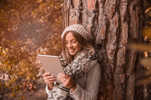 Mujer alegre que usa la tableta cerca del árbol