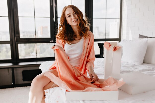 Mujer alegre en ropa de dormir sonriendo en el dormitorio. Chica rizada interesada sentada en la cama con expresión de cara feliz.