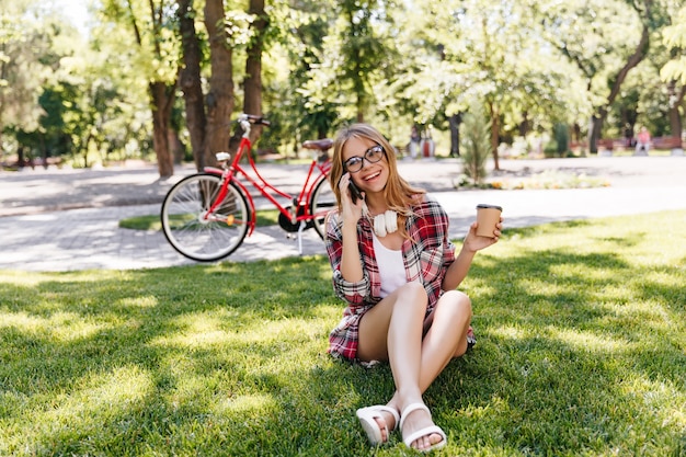 Mujer alegre en ropa de verano sentada en la hierba y tomando café. Tiro al aire libre de fascinante chica con gafas hablando por teléfono sobre la naturaleza.