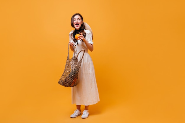 Mujer alegre saca naranja de la bolsa de hilo. Señora en vestido midi blanco posando sobre fondo naranja.