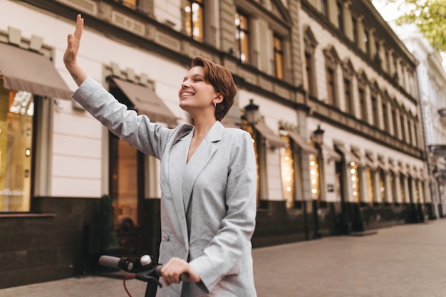 Mujer alegre saludando con la mano mientras camina. Chica guapa en traje gris montando scooter eléctrico y sonriendo afuera