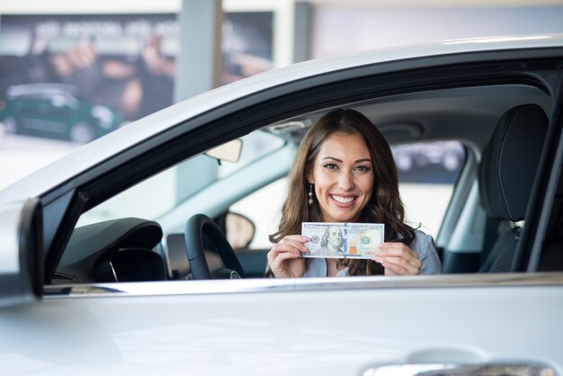 Mujer alegre sentada en el coche nuevo con billete de dólar estadounidense