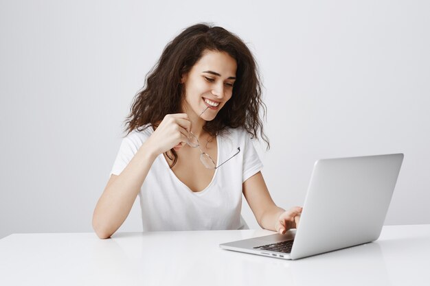 Mujer alegre sonriendo complacido en portátil mientras sentarse escritorio